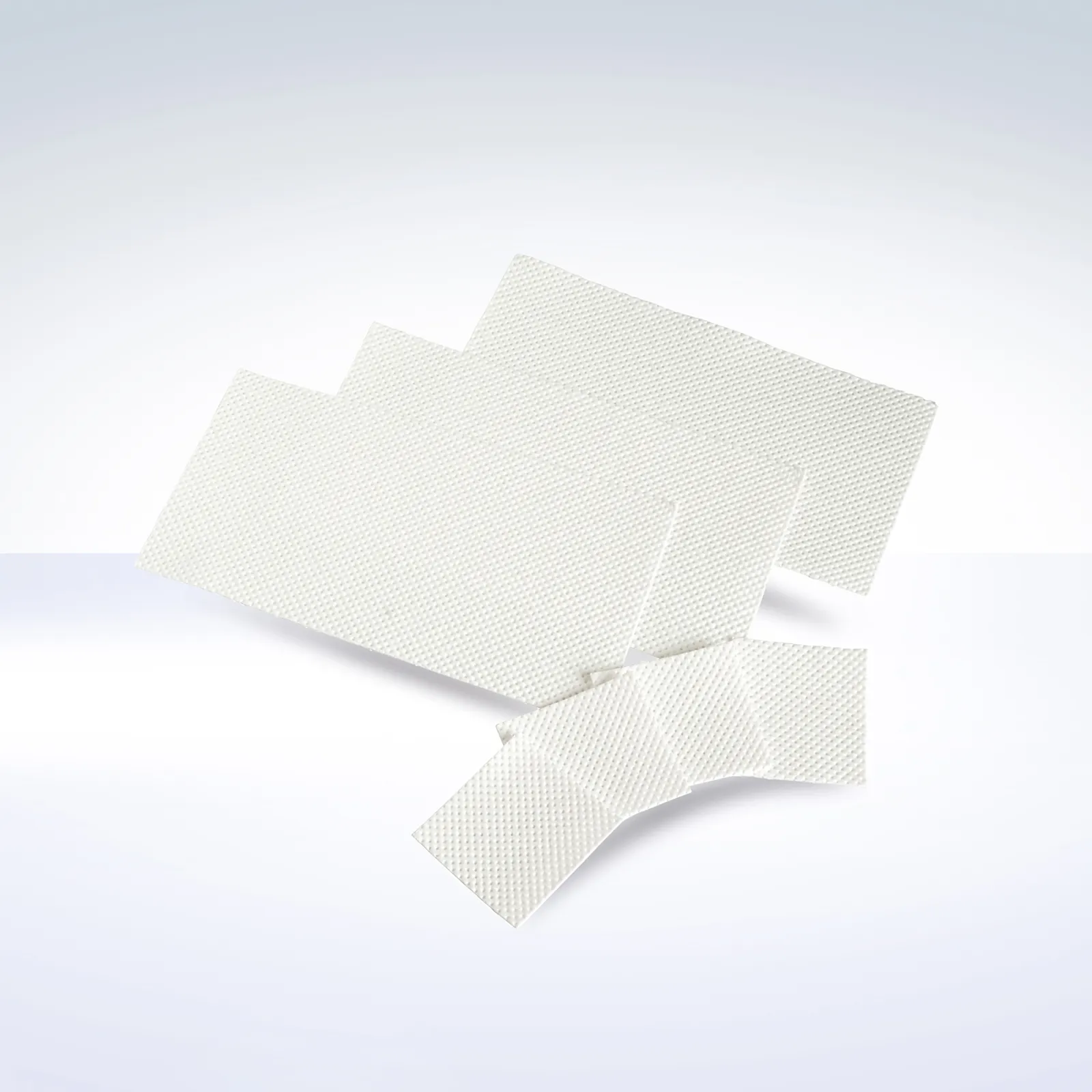 Several white DEBAMED® absorbent pads in different rectangular sizes arranged in a fan-like spread on a soft white-to-light-blue gradient background. The pads have a fine dimpled surface texture.